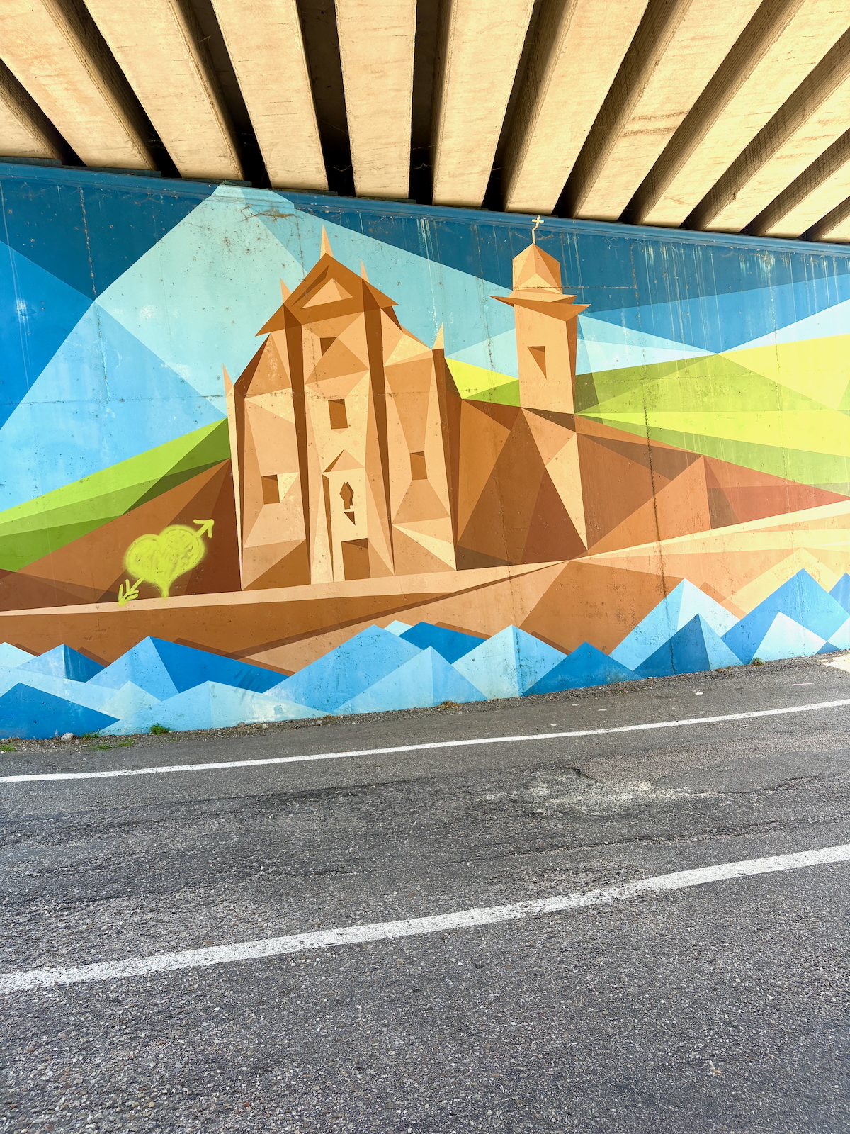 Geometric mural on a wall under a bridge, showing a stylized church with brown angular shapes, blue sky, and rolling hills in greens and yellows, beside a paved road.