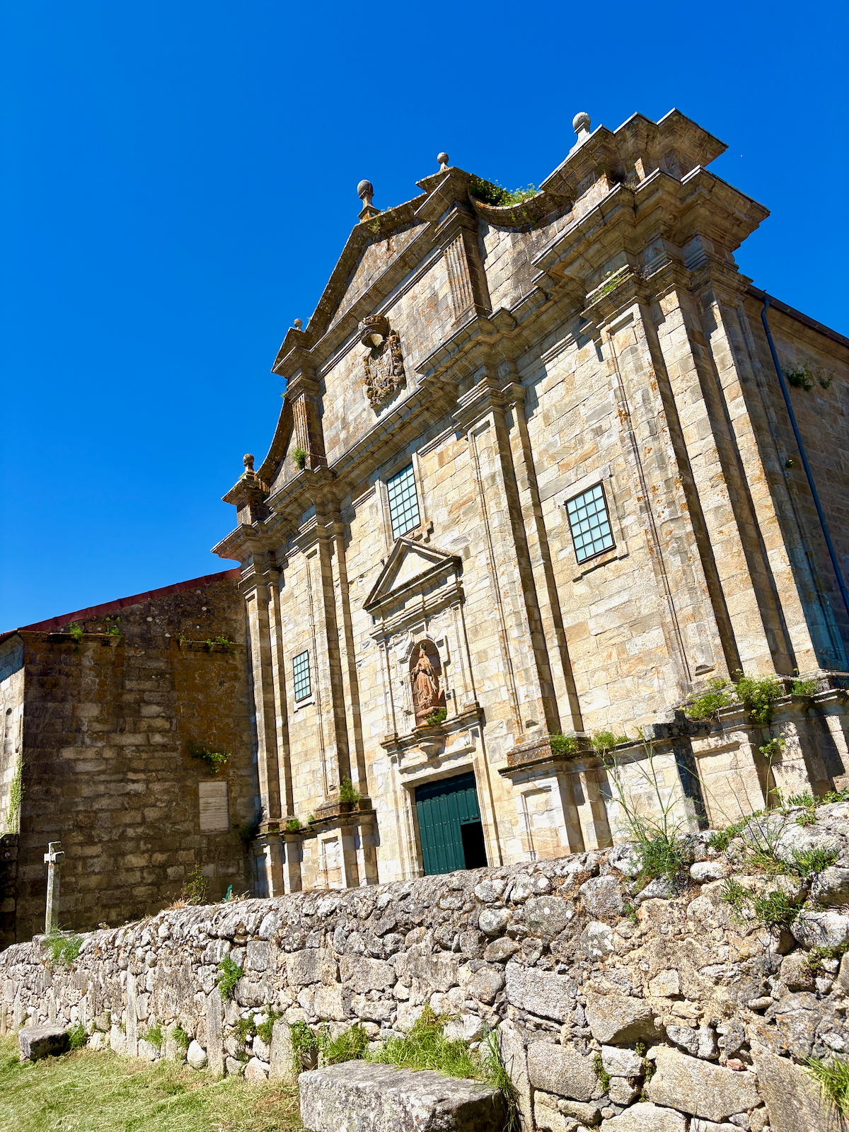 Historic stone church facade with ornate pediment and statue niche against a bright blue sky; foreground stone wall and greenery.