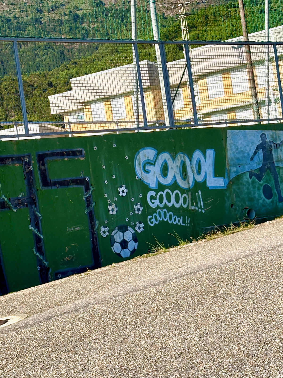 Green barrier wall painted with soccer-themed graffiti, featuring the word 'GOOL' and several soccer ball drawings beside a paved path.