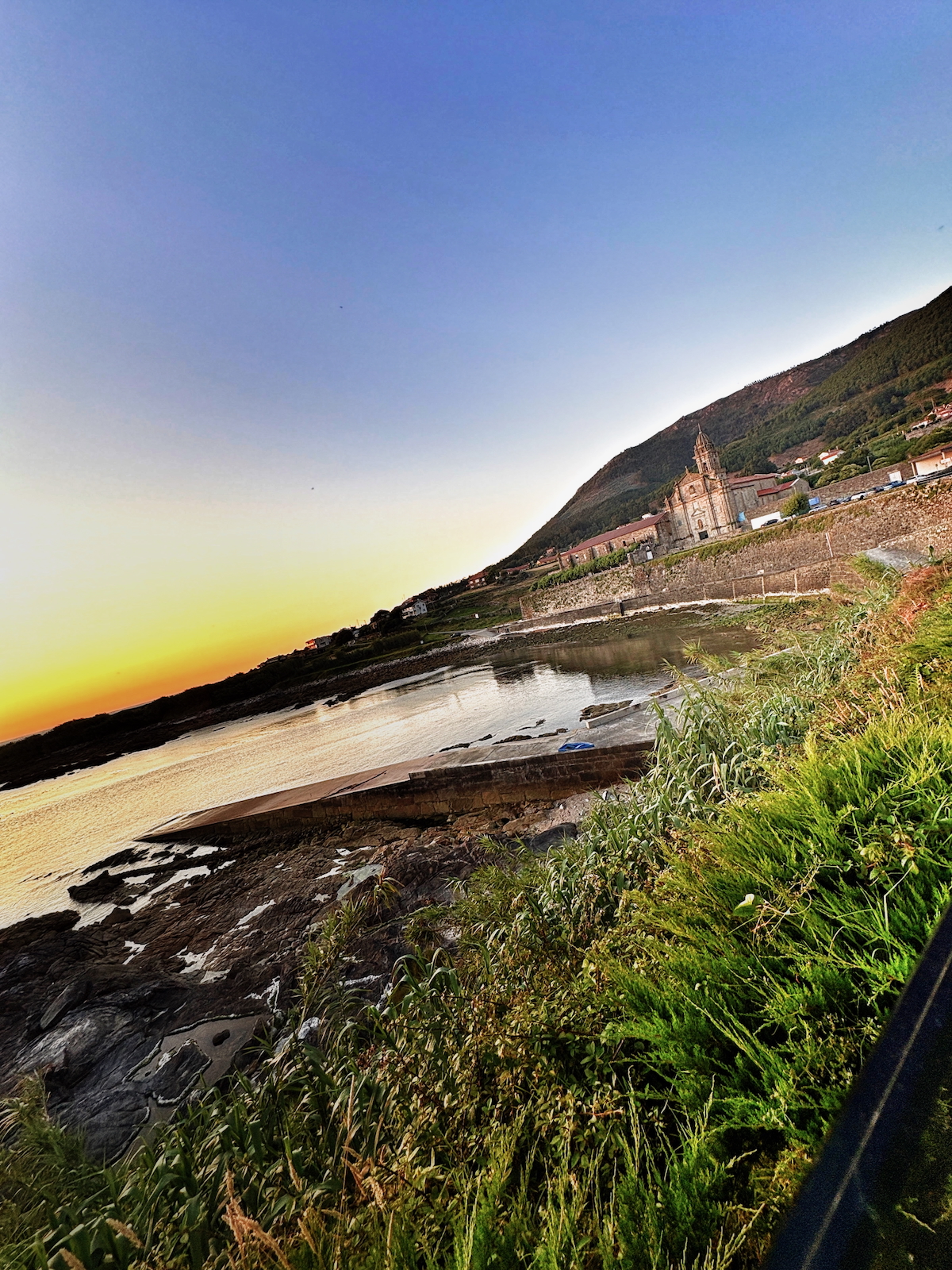 Sunset over a coastal town with a church on a hill, calm water reflecting the sky, and grassy foreground by the shoreline