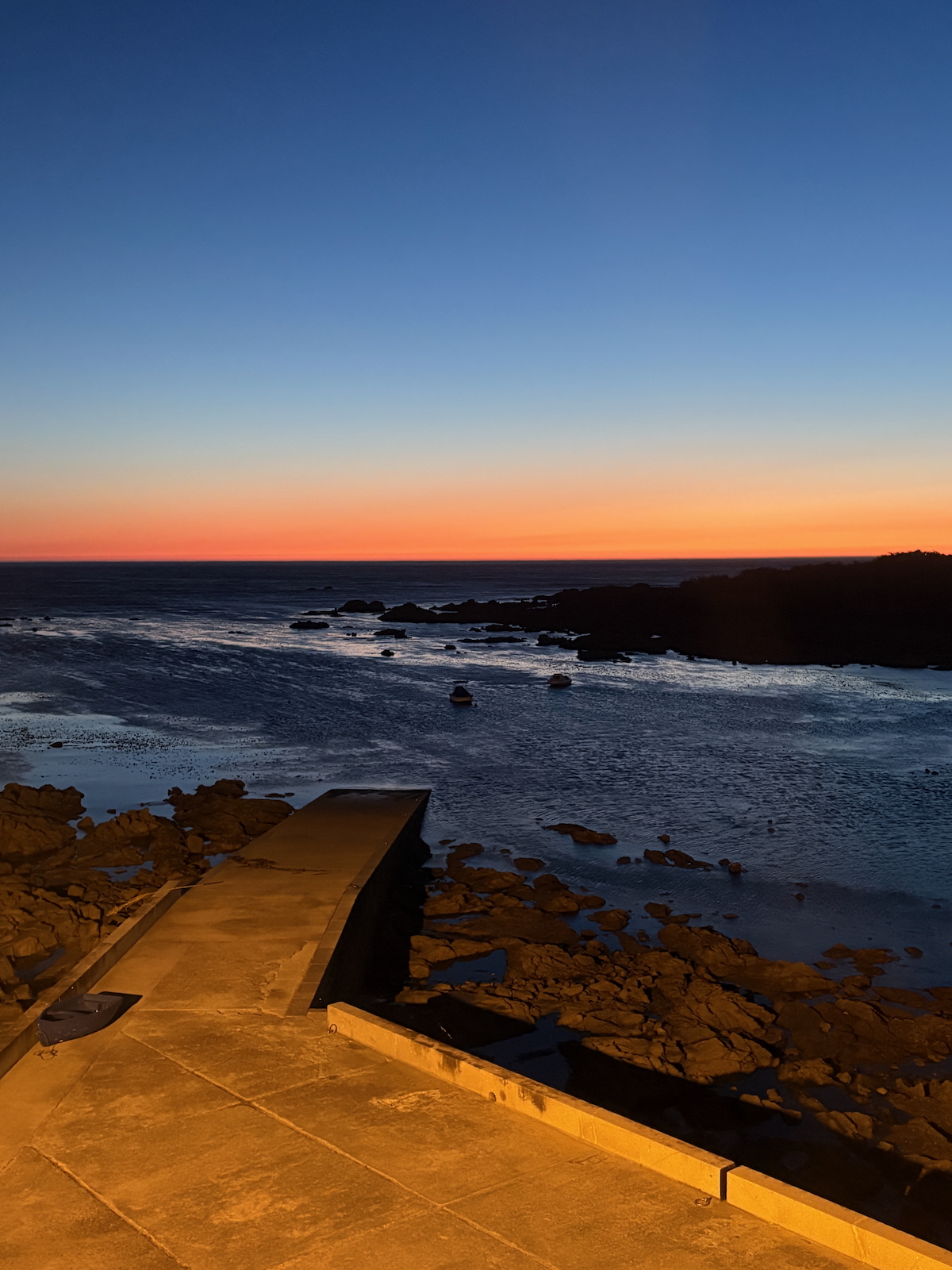 Sunset over the ocean with a gradient orange-to-blue sky, rocky shoreline, and a concrete pier leading toward calm waters.